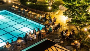 Un área de comedor al aire libre con mesas y sillas dispuestas alrededor de una piscina, rodeada de follaje y árboles.