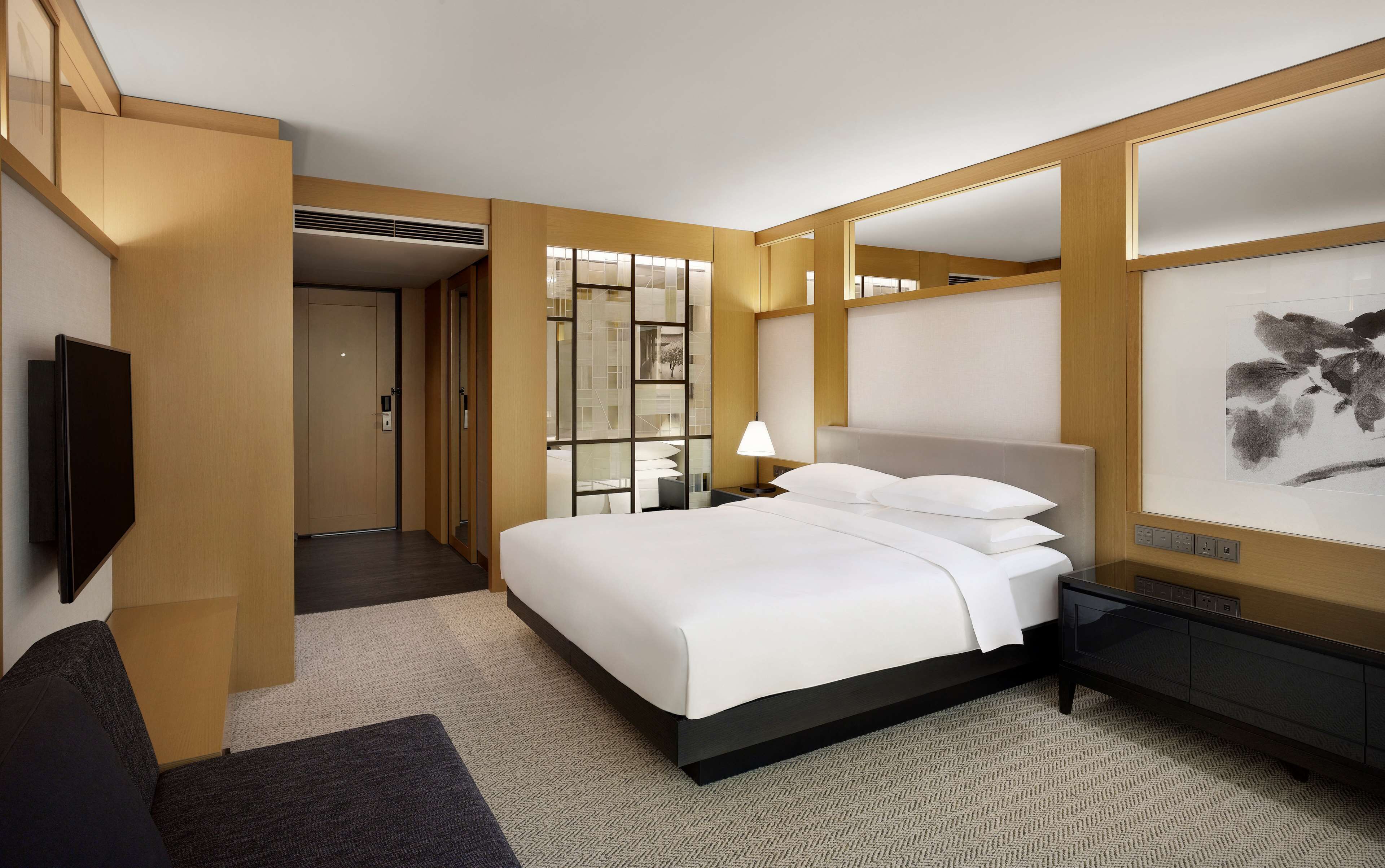 A modern hotel room with a large bed, a bedside table with a lamp, a TV mounted on the wall, and a large artwork on the wall.