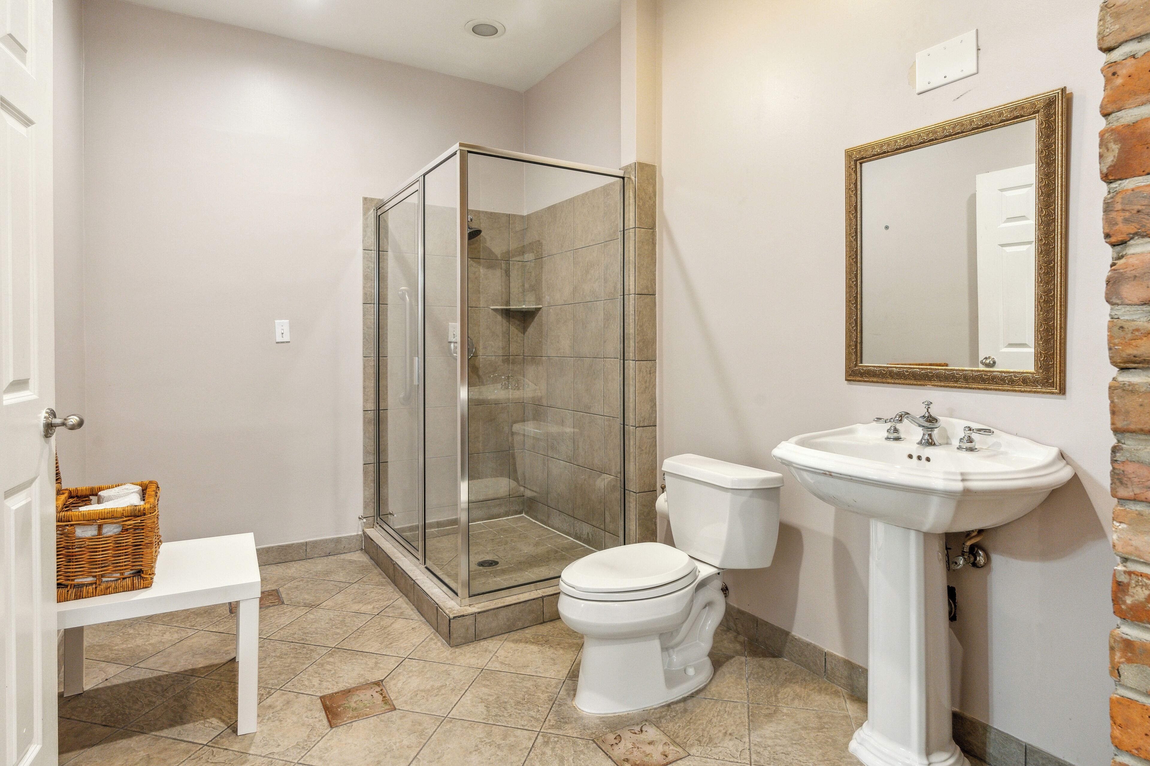 A bathroom with a glass-enclosed shower, a white toilet, a white sink, and a wicker basket on a white bench.