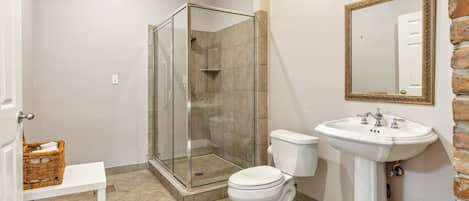 A bathroom with a glass-enclosed shower, a white toilet, a white sink, and a wicker basket on a white bench.
