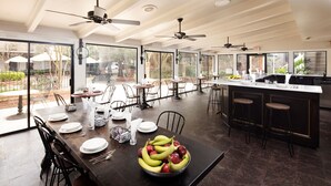 Uma sala de jantar espaçosa com uma mesa comprida, cadeiras e uma cesta de frutas. Há ventiladores de teto e janelas amplas que proporcionam luz natural.