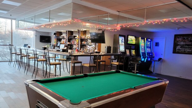A pool table in a room with a bar area, chairs, and arcade games.