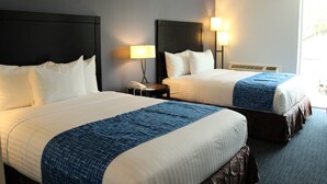 A hotel room with two beds, each with white linens and a blue accent. There are bedside lamps and a phone on a nightstand. - Travelodge by Wyndham Water’s Edge Hotel - Racine (Racine)