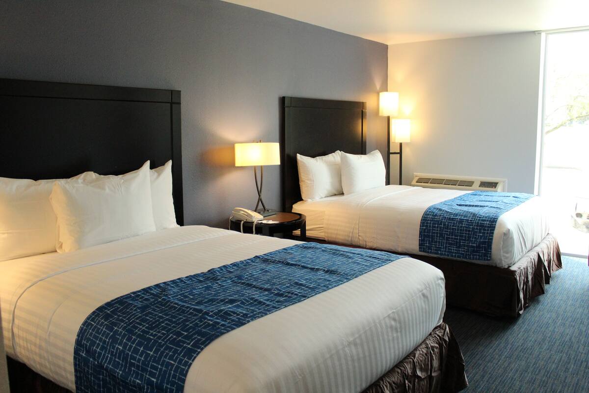 A hotel room with two beds, each with white linens and a blue accent. There are bedside lamps and a phone on a nightstand.