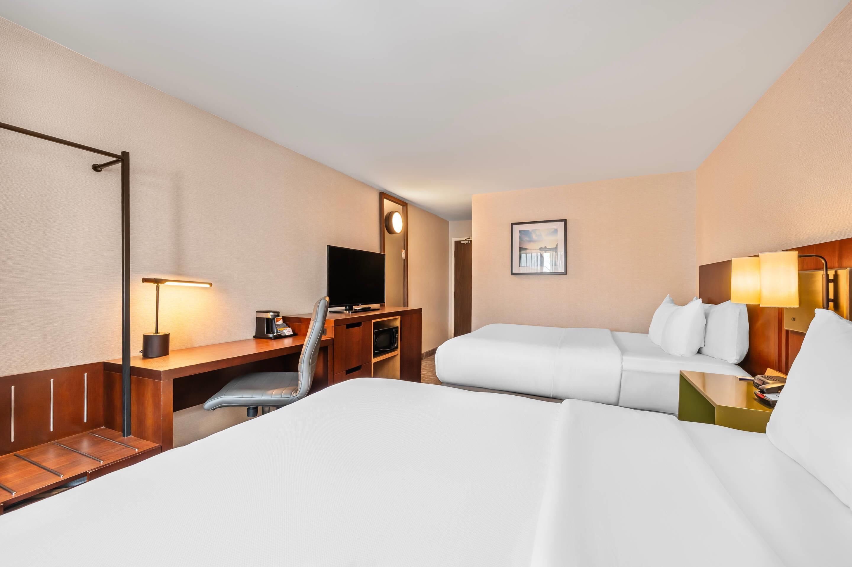 A hotel room with two beds, a desk with a flat-screen TV, a chair, a desk lamp, a bedside table with a lamp, and a framed picture on the wall.