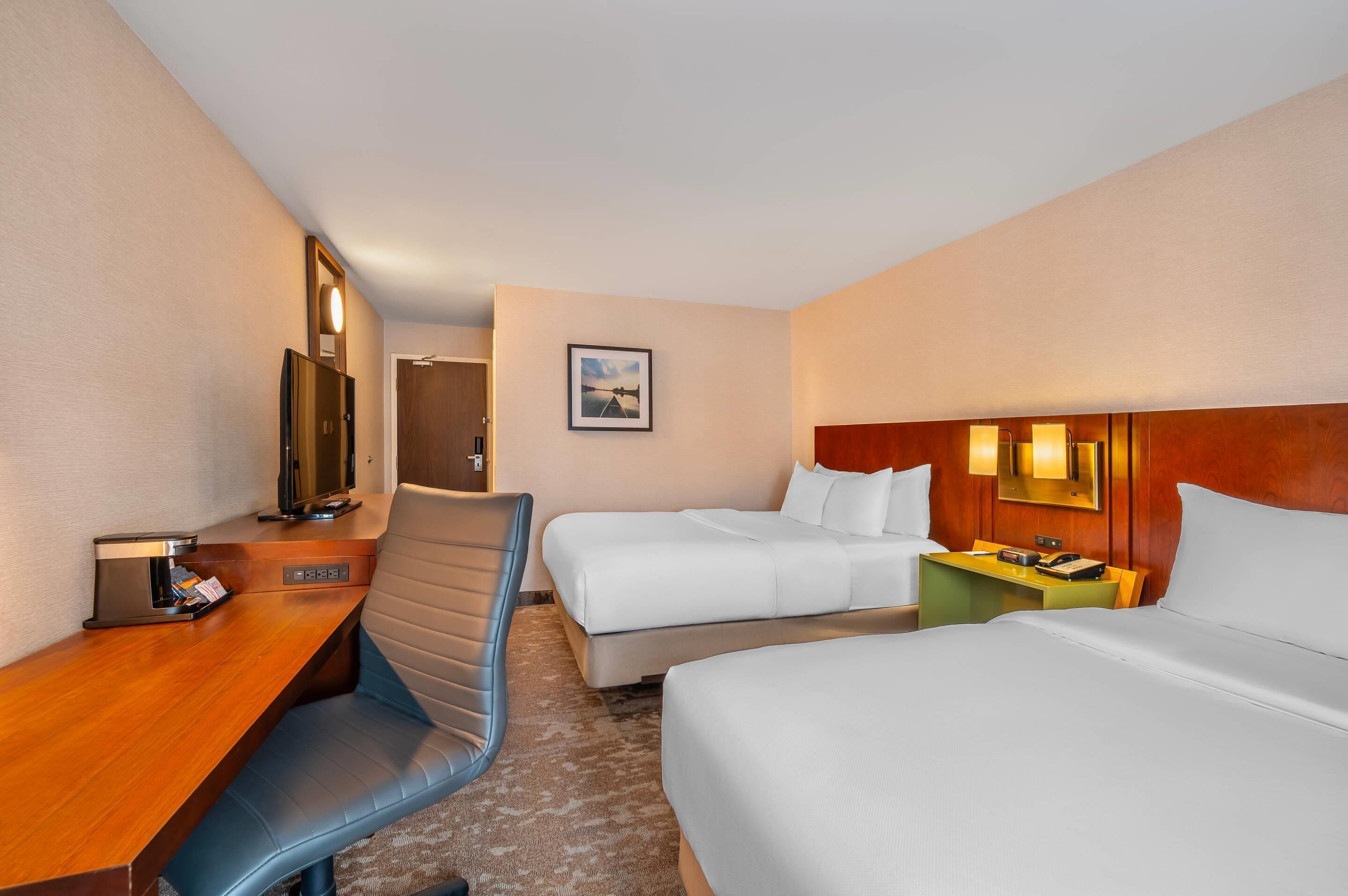 A hotel room with two beds, a desk, a chair, a mirror, a picture on the wall, and a wooden headboard.