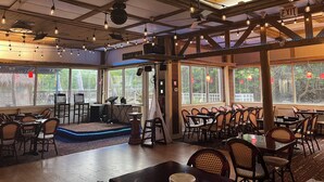 Ein Restaurant mit Holzbalken, Lichterketten und großen Fenstern. Tische und Stühle sind zum Essen bereitgestellt.