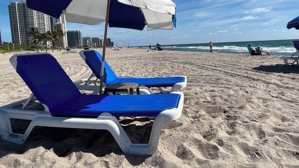 Ein Strand mit zwei blauen Liegestühlen unter einem weiß-blauen Sonnenschirm, feinem Sand und einer Stadtkulisse im Hintergrund.