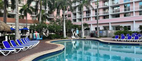 Un espace piscine d’hôtel avec des chaises longues, des palmiers et un immeuble à plusieurs étages doté de balcons.