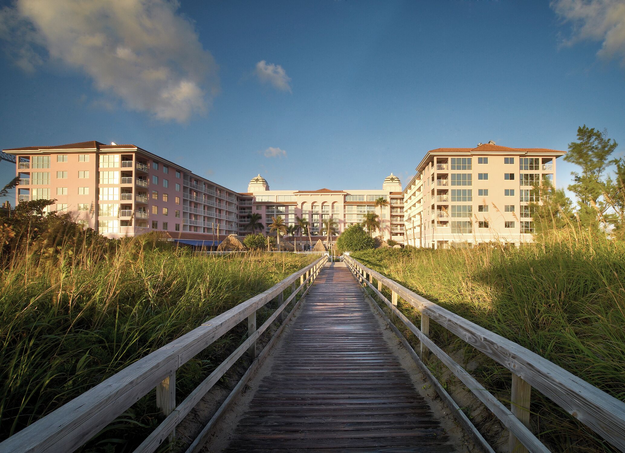 A wooden boardwalk leading to a hotel with multiple floors and balconies, surrounded by tall grass and trees.