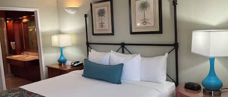 Habitación de hotel con cama, dos lámparas fijadas a la pared, una mesita de noche, un tocador, baño con lavamanos y espejo, y dos cuadros enmarcados en la pared.