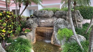 Ein angelegter Garten mit Wasserspiel, Palmen und einem rosafarbenen Gebäude im Hintergrund.