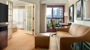 A hotel room with a large bed, a television, a sofa, an ottoman, a desk, and a balcony with a chair.