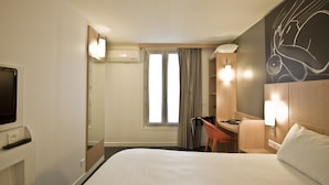 Habitación de hotel con cama, escritorio, silla, televisión, ventana con cortinas y una obra de arte en la pared.