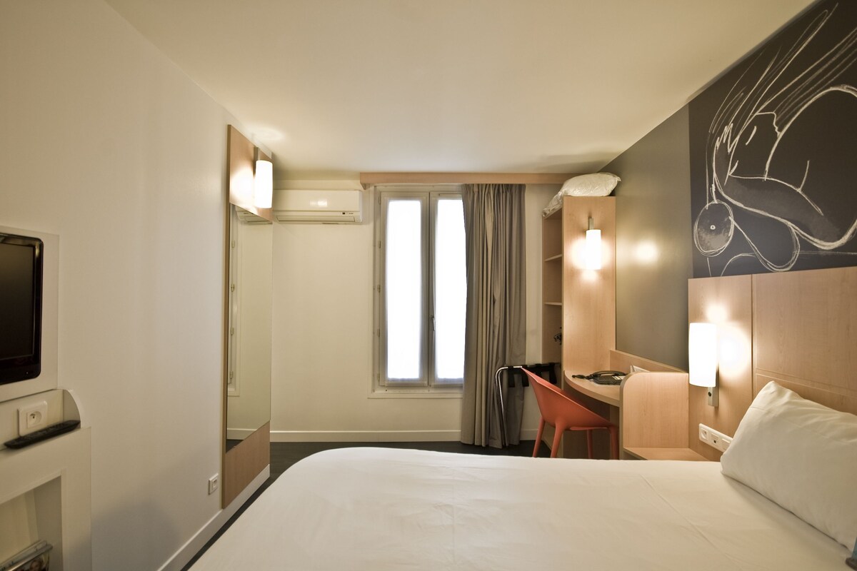 Habitación de hotel con cama, escritorio, silla, televisión, ventana con cortinas y una obra de arte en la pared.