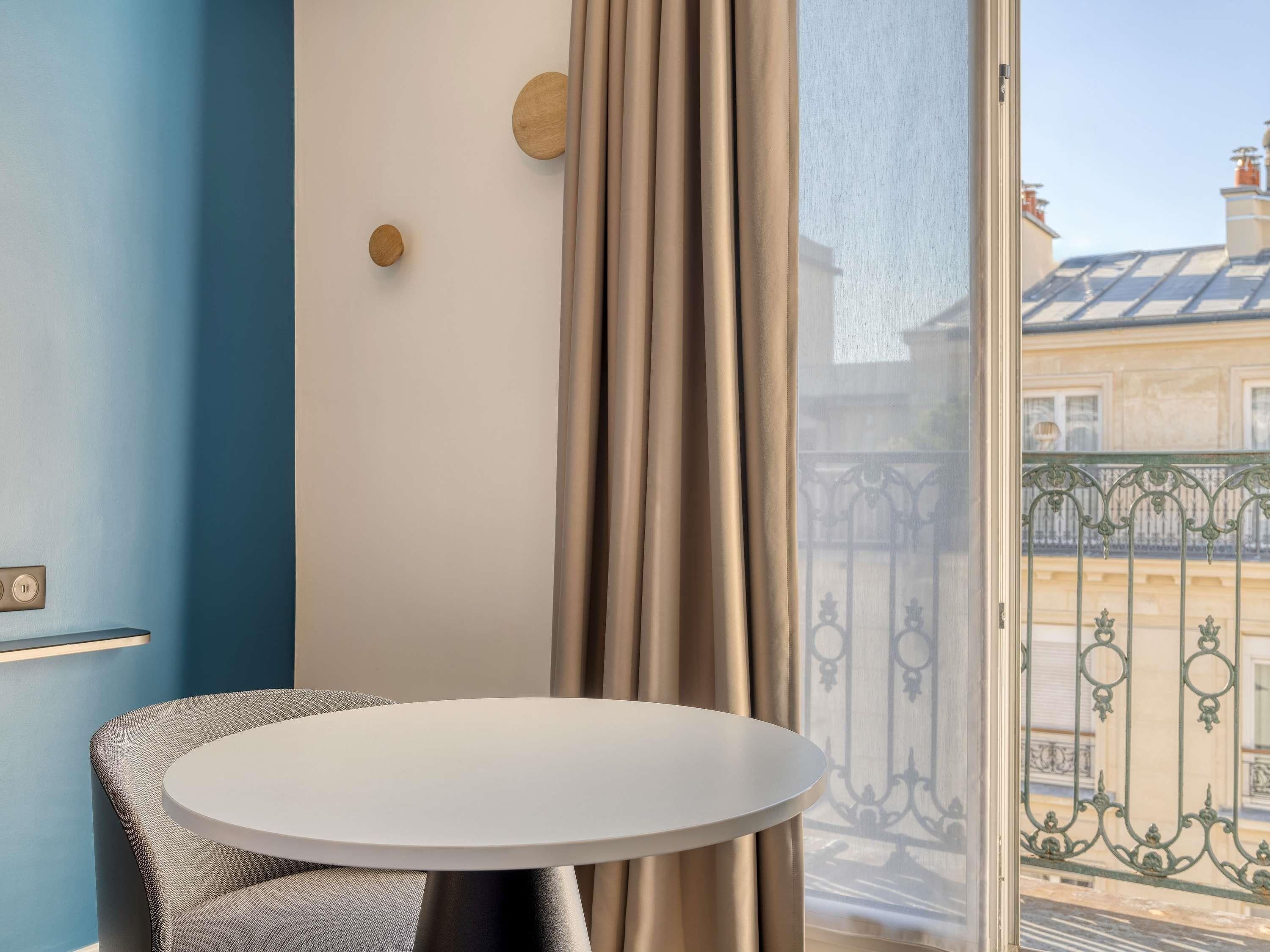 Una habitación con una mesa redonda, una silla, una ventana con cortinas y vistas a edificios.