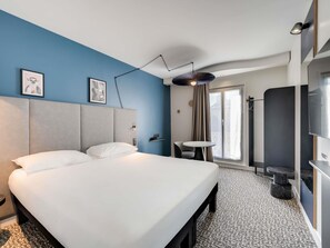 A hotel room with a large bed, a small table, a chair, a TV, and a bathroom with a shower. - ibis Paris Père Lachaise (Paris)