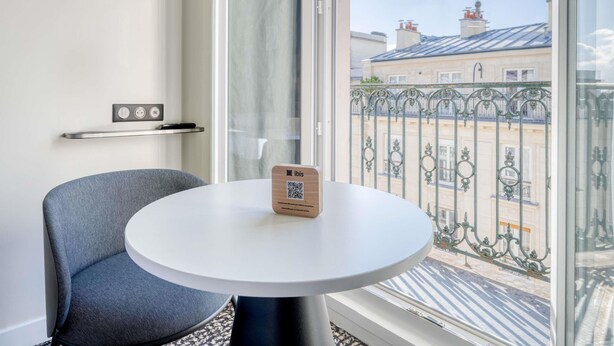 A hotel room with a round white table, a grey chair, a wooden door, and a window with a view of buildings and a metal fence. - ibis Paris Père Lachaise (Paris)