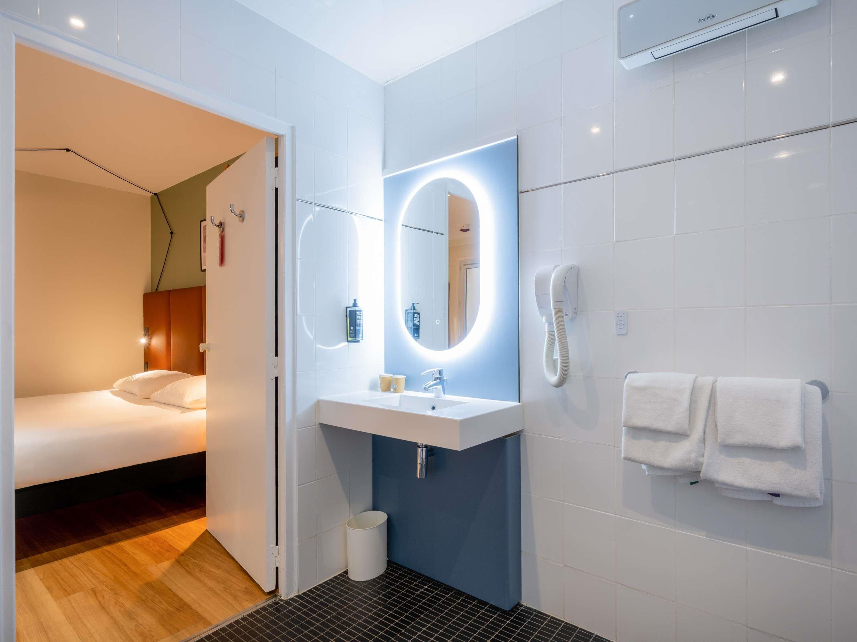 A modern hotel room with a bed, a bathroom with a sink, mirror, and a telephone.