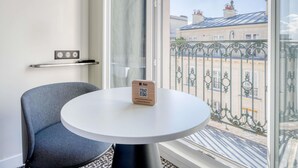 A hotel room with a round white table, a grey chair, a wooden door, and a window with a view of buildings and a metal fence. - ibis Paris Père Lachaise (Paris)