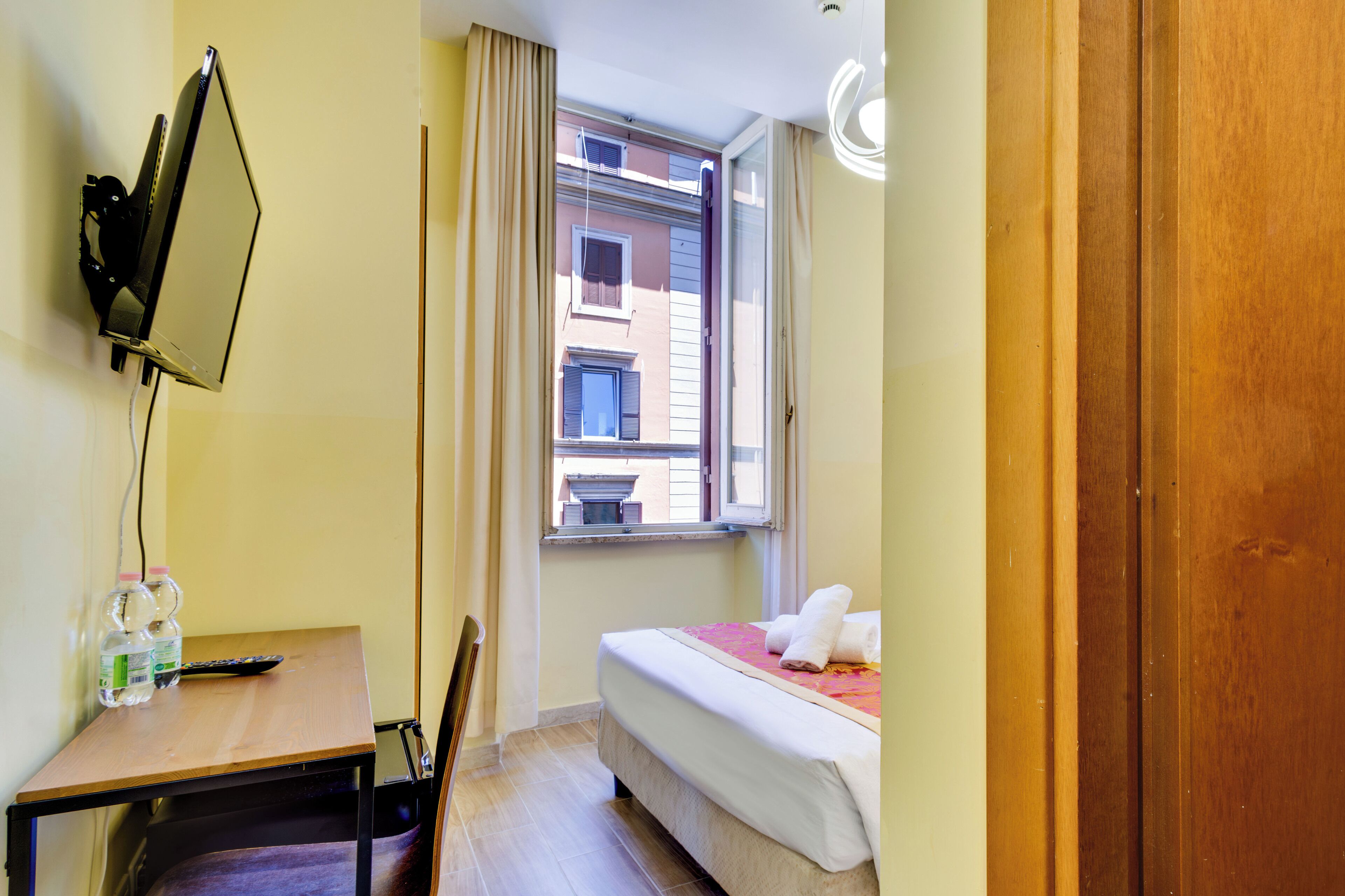 A hotel room with a bed, a desk with a chair, a window with curtains, and a wall-mounted mirror.