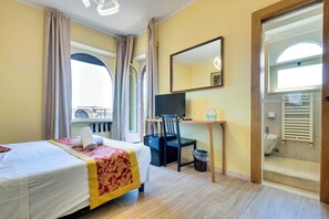 A hotel room with a bed, desk, chair, and a view of the city through large windows.