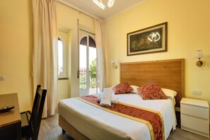 A hotel room with a large bed, a wooden headboard, a painting on the wall, a bedside table with a lamp, and a view through an open door.