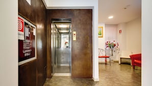An elevator in a hallway with a wooden wall and a red sign on the left. There is a white chair and a red chair in the room. - Hotel Terminal (Rome)