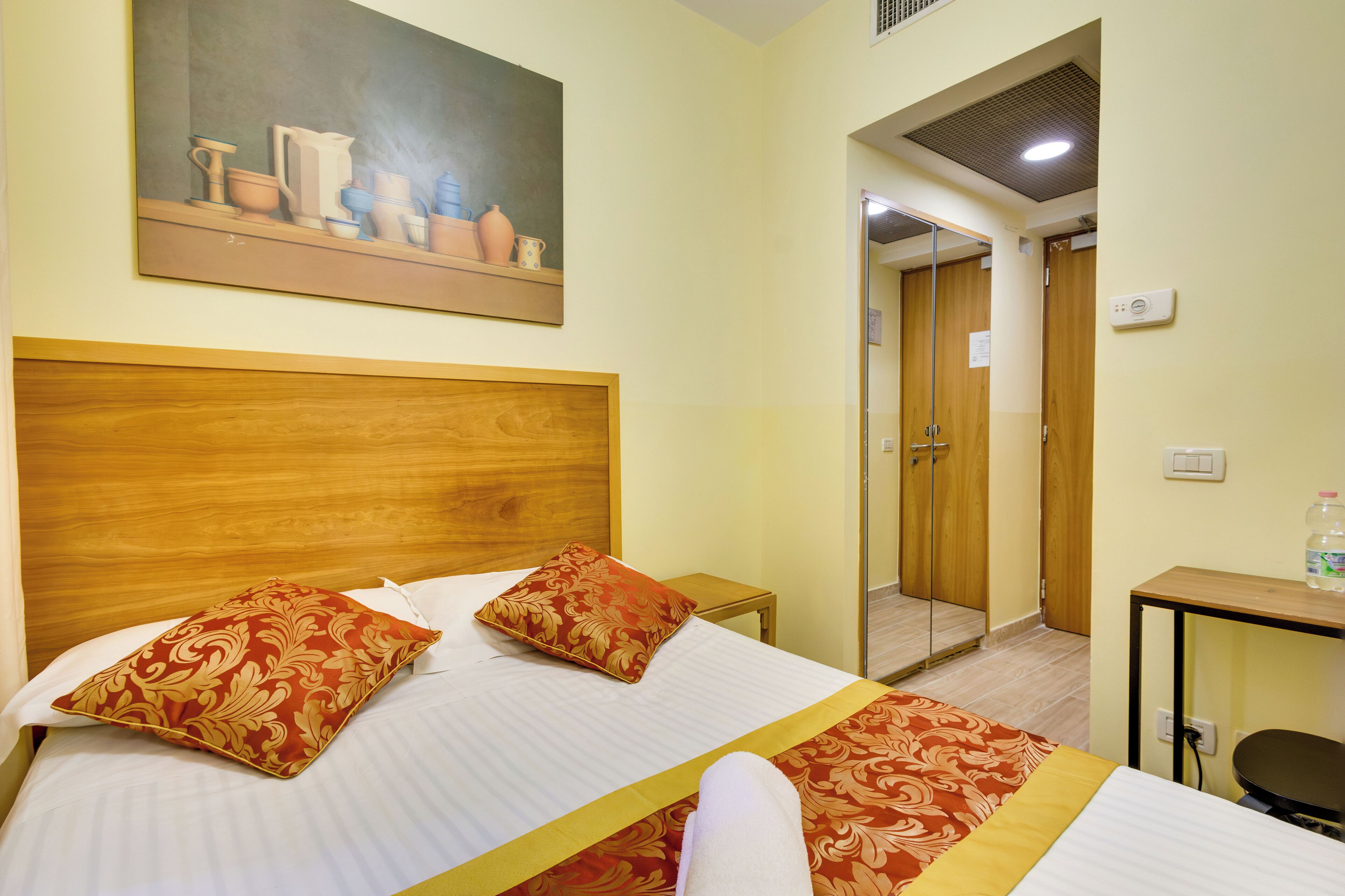 Quarto de hotel com cabeceira de madeira, duas almofadas decorativas, cama com colcha listrada, um quadro sobre a cama, uma mesinha com uma garrafa e uma porta que dá para o banheiro.