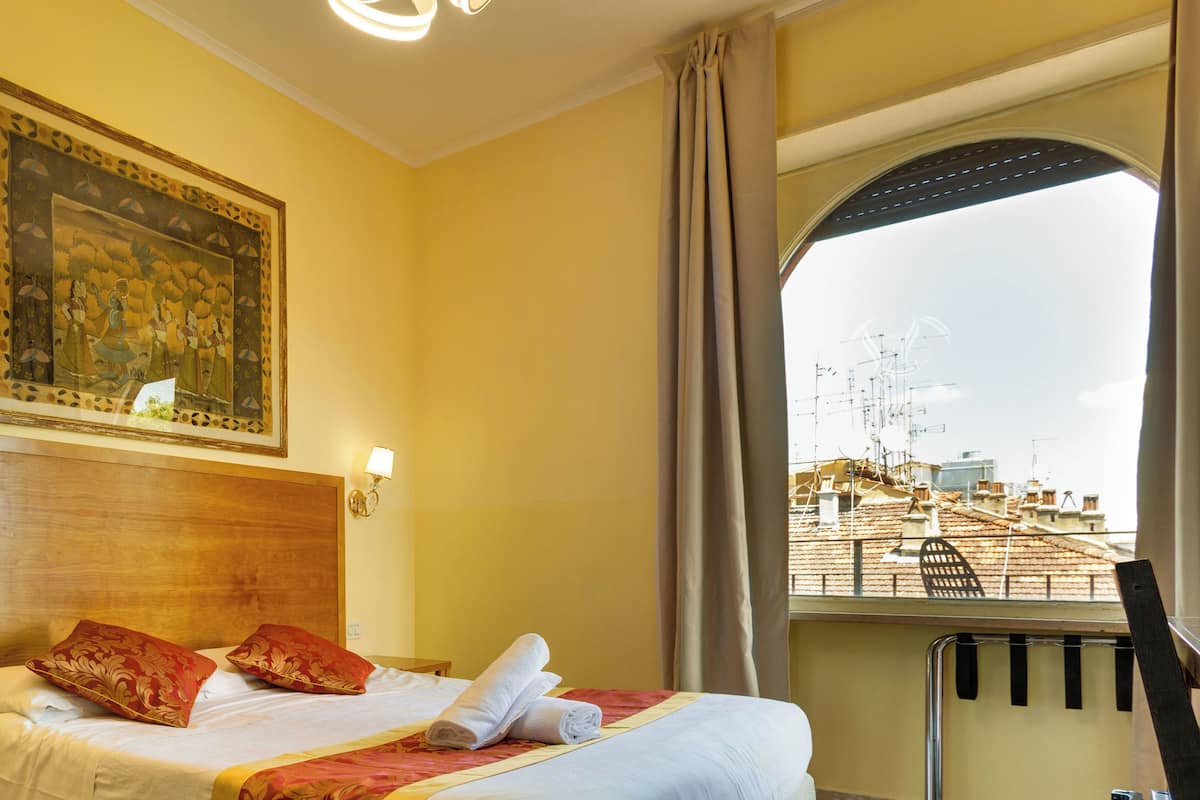 Une chambre d’hôtel avec un lit, un tableau, une fenêtre donnant sur des immeubles et une chaise près de la fenêtre.