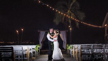 A wedding ceremony with a couple kissing under string lights, surrounded by white chairs and a backdrop with drapes.