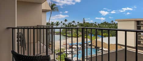 A balcony with wicker chairs, a pool area with umbrellas, and a view of palm trees and a building.