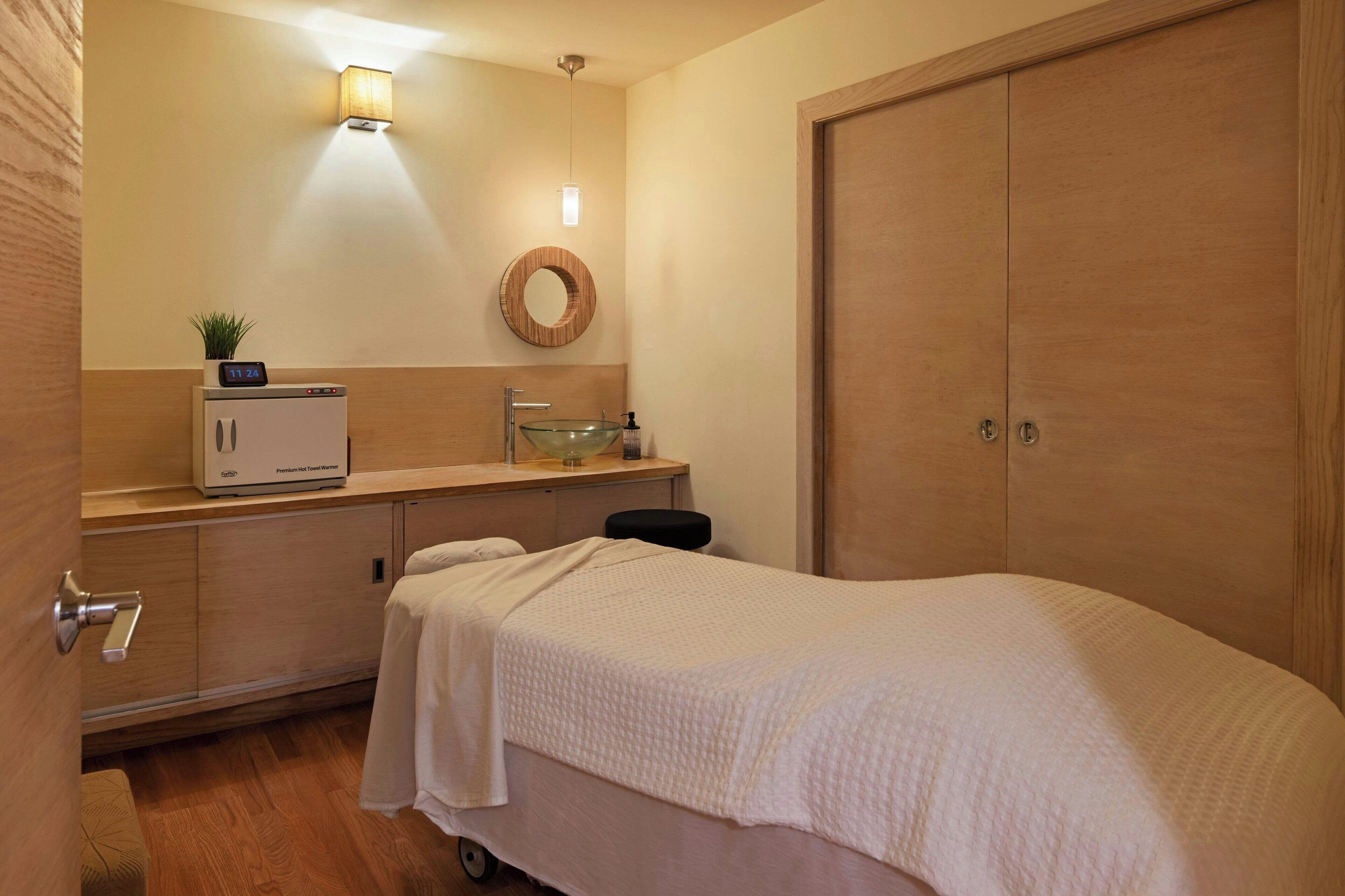 A hotel room with a bed, a bedside table with a lamp, a small plant, a round mirror, and a wardrobe.