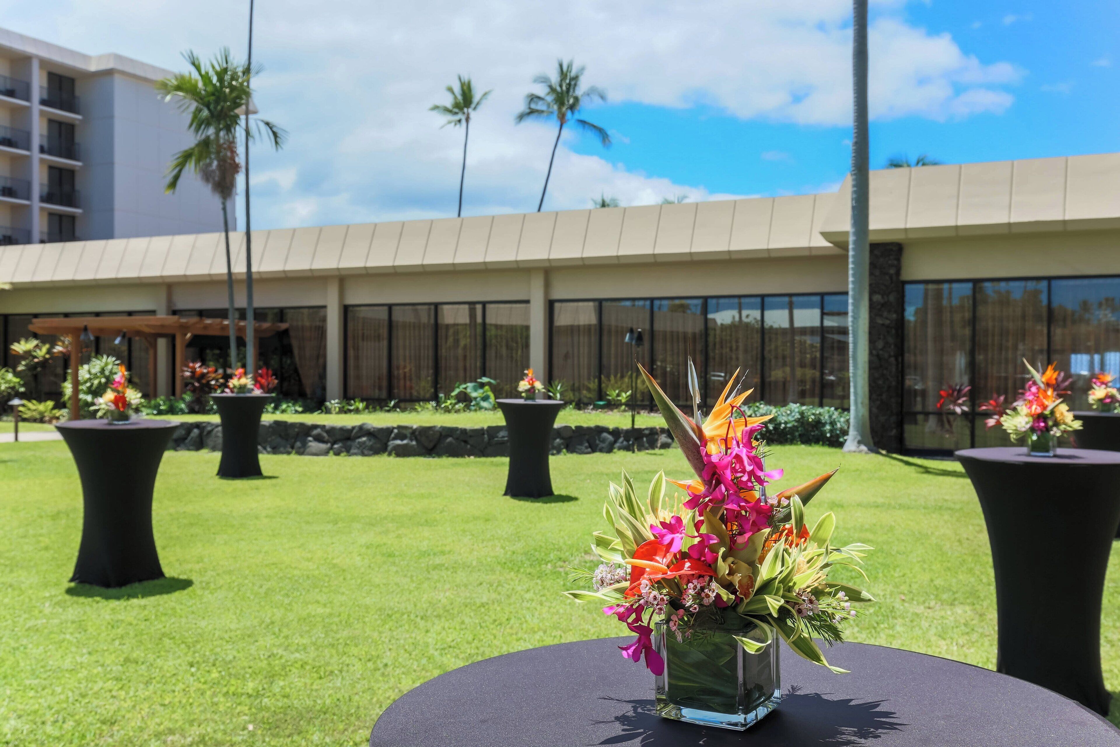 Un patio de hotel con mesas al aire libre, arreglos florales y un edificio de techo plano y ventanas grandes.