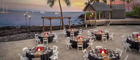 An outdoor dining area with round tables and white chairs, set against a backdrop of a sunset over the ocean with boats and palm trees.