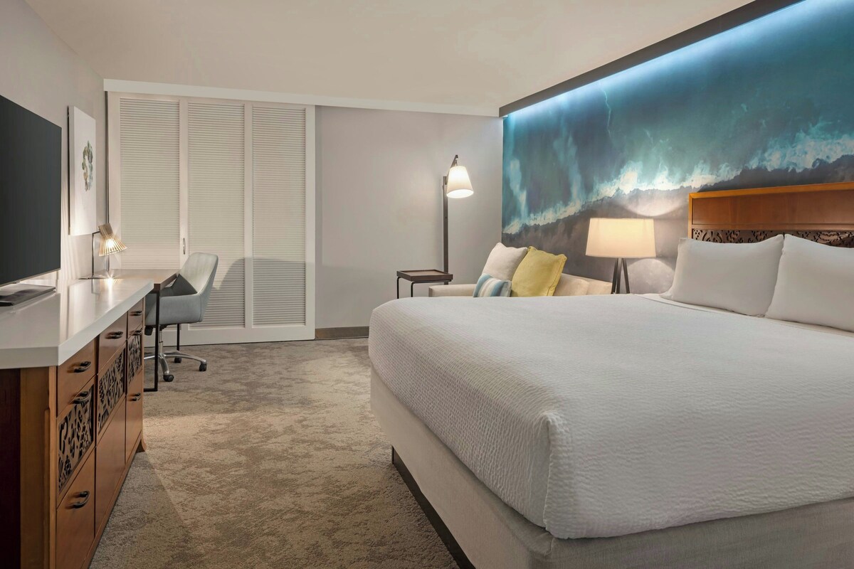 Una camera d'hotel con un letto grande, una scrivania con una sedia, una TV, un quadro raffigurante un mare in tempesta e una lampada.