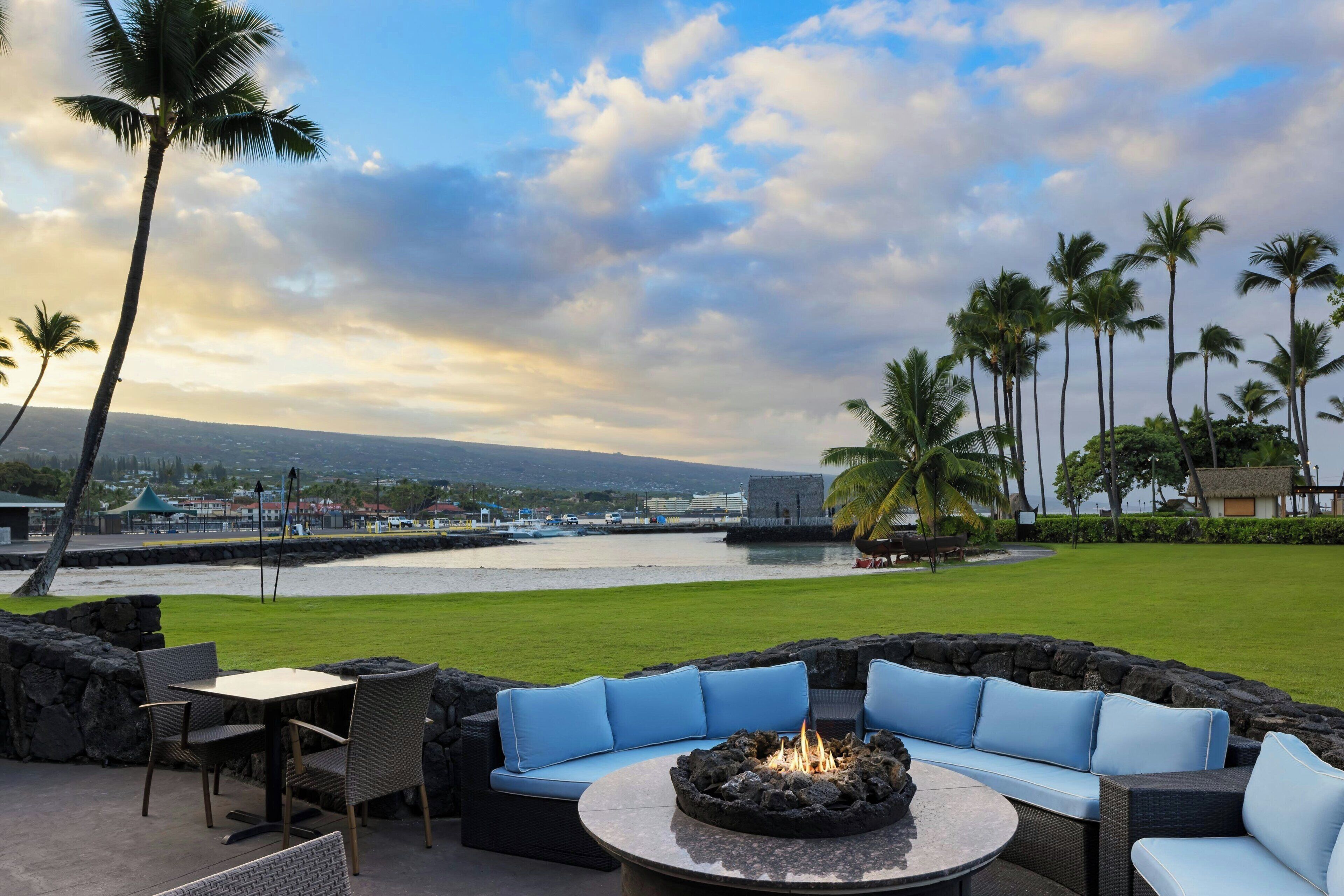 An outdoor seating area with a fire pit, overlooking a body of water and palm trees.