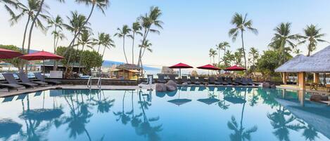 A pool with clear water, surrounded by lounge chairs and umbrellas, palm trees, and a thatched-roof structure.