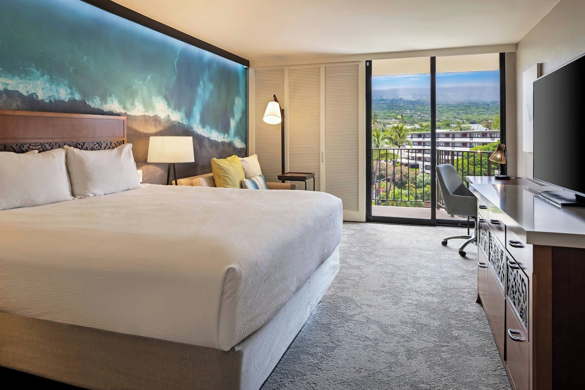 Une chambre d’hôtel avec un grand lit, un bureau, une chaise, une télévision et une vue sur la ville grâce à de grandes fenêtres.