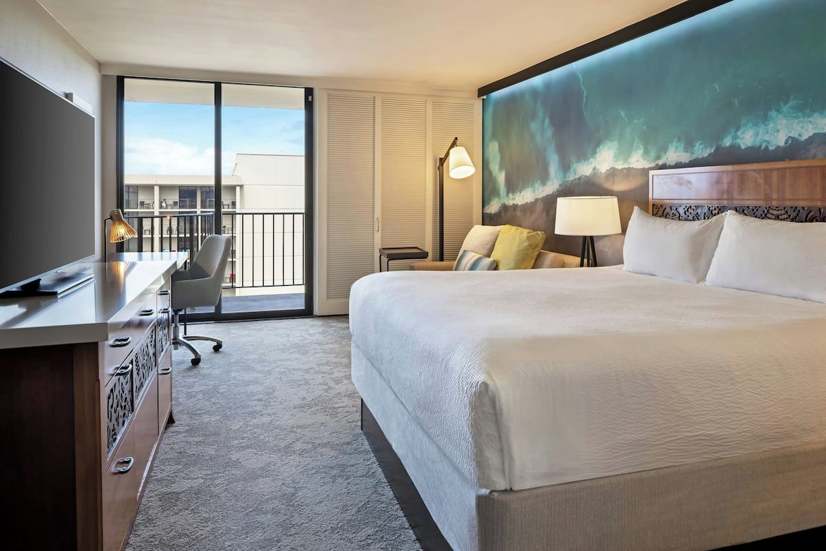 Une chambre d’hôtel avec un grand lit, un bureau avec une chaise, un ordinateur portable et une vue sur l’extérieur grâce à une porte coulissante.