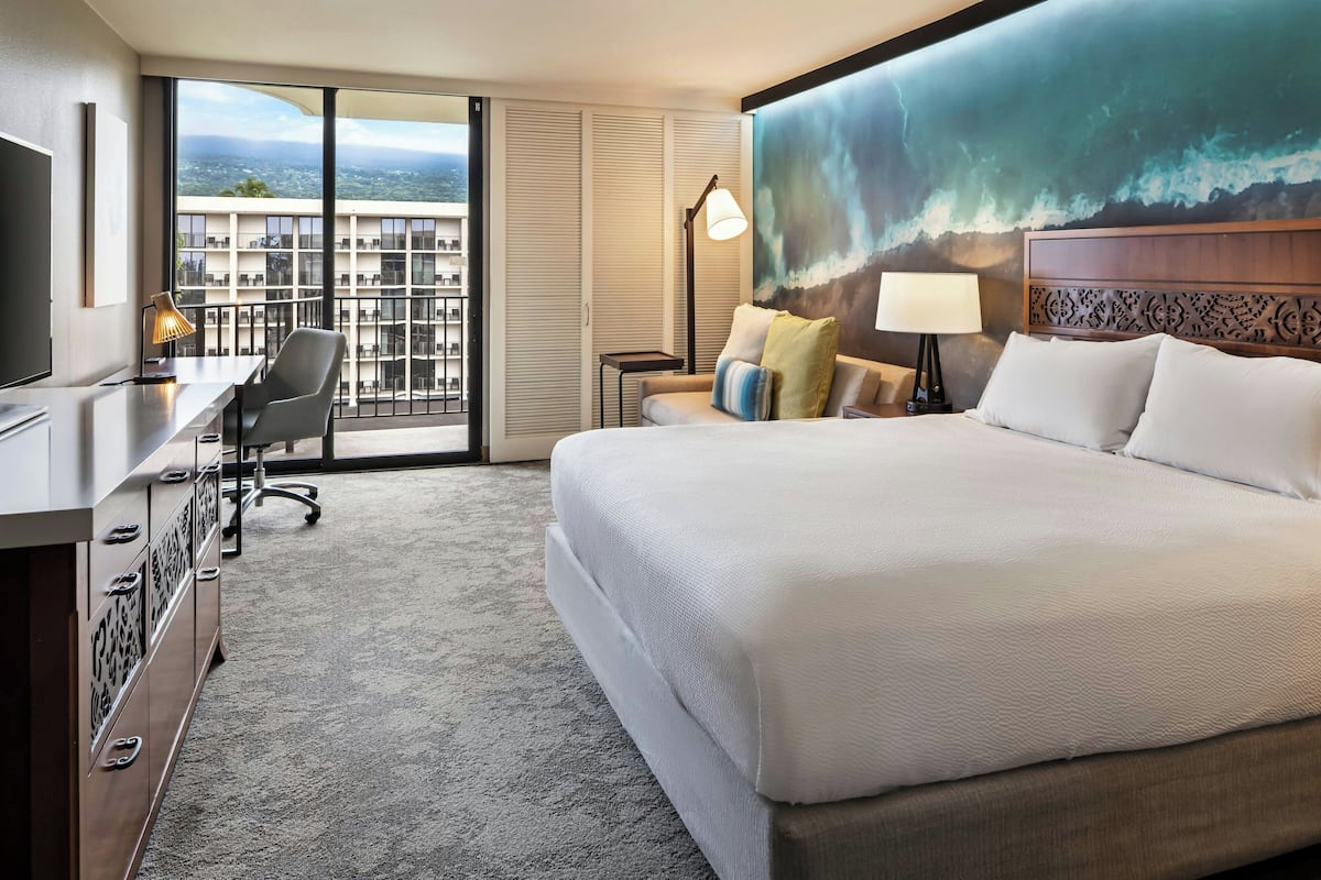 Une chambre d’hôtel avec un grand lit, un bureau avec une chaise, une vue sur la ville grâce à de grandes fenêtres et un tableau représentant une vague accroché au mur.