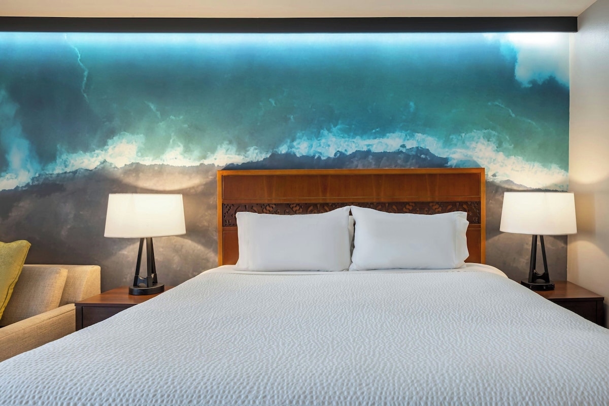 Hotelkamer met een groot bed, twee bedlampen, een houten hoofdeinde en een muurschildering van een berglandschap.