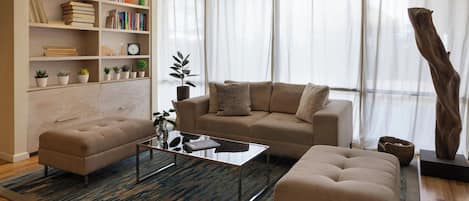 A modern living room with a sofa, ottoman, and a coffee table. There is a bookshelf with books and decorative items.