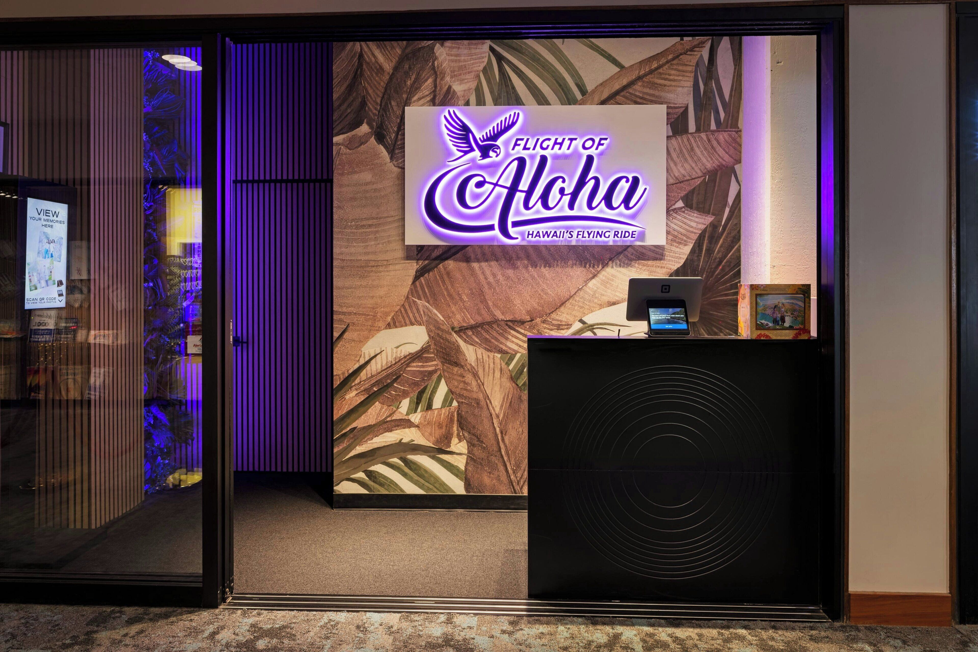 Un espace d’accueil avec une enseigne indiquant « FLIGHT OF ALOHA HAWAII’S FLYING RIDE » et un écran d’ordinateur posé sur un bureau.
