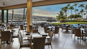 An outdoor dining area with wicker chairs and tables, large windows offering a view of palm trees and a body of water. - Courtyard by Marriott King Kamehameha's Kona Beach Hotel (Kailua-Kona)