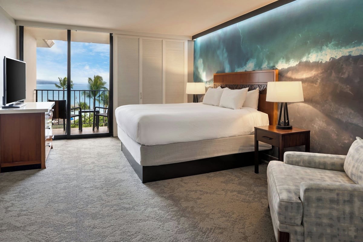 Une chambre d’hôtel avec un grand lit, un bureau avec une télévision, une chaise, un balcon avec vue sur l’océan et un tableau au mur.
