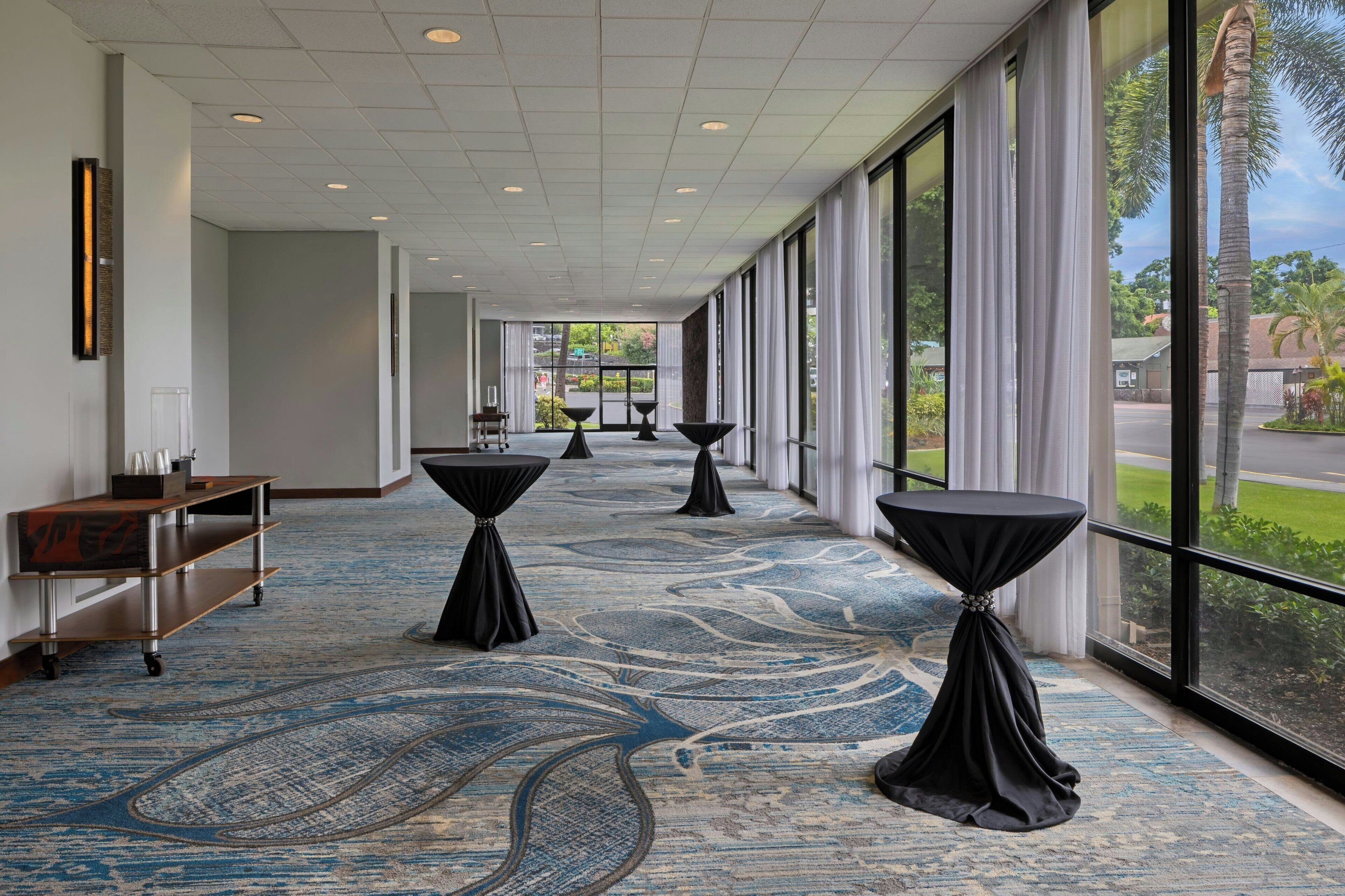 Un couloir d’hôtel avec un tapis à motifs, de grandes fenêtres et des tables recouvertes de nappes noires.