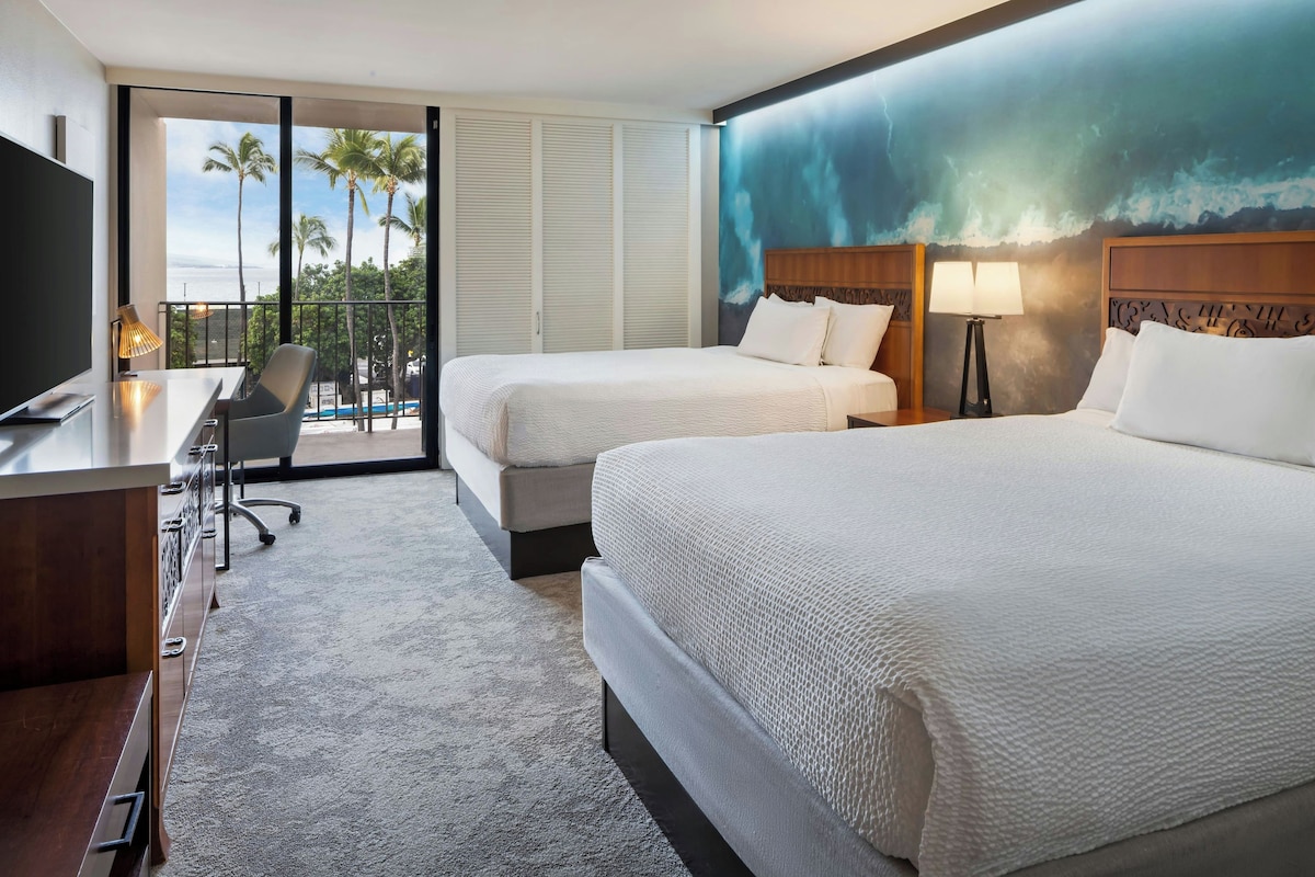 Een hotelkamer met twee bedden, een bureau, een stoel, een televisie en uitzicht op palmbomen en de oceaan.