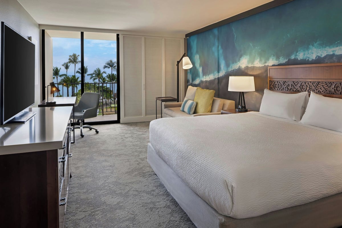 Une chambre d’hôtel avec un grand lit, un bureau avec une télévision, une chaise, une lampe et une vue sur les palmiers et l’océan, grâce à de grandes fenêtres.