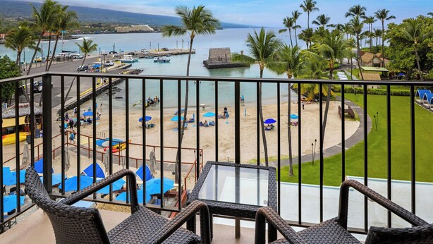 A balcony with wicker chairs overlooking a beach area with umbrellas and a dock with boats. - Courtyard by Marriott King Kamehameha's Kona Beach Hotel (Kailua-Kona)